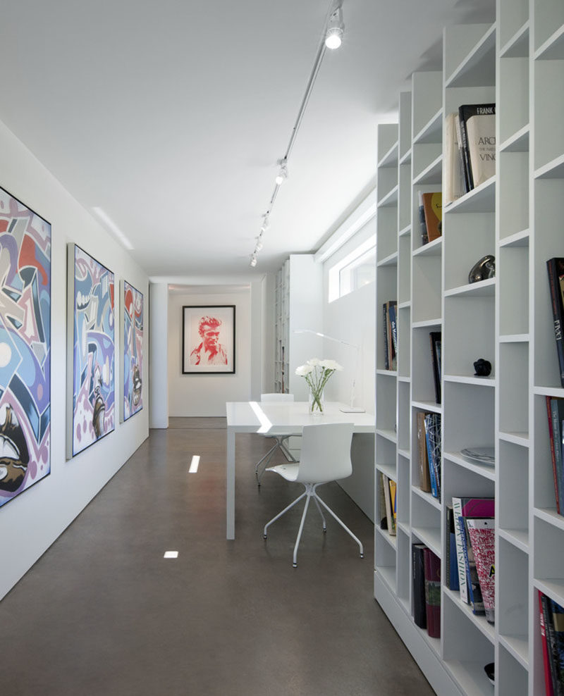 This bright white hallway, also known as the home's library, is filled with colorful artwork, floor-to-ceiling bookshelves, and a small desk area.