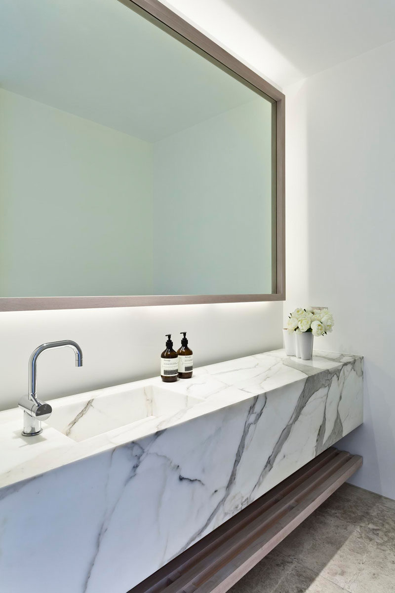 This modern bathroom has a backlit mirror, a marble vanity and an open wooden shelf for additional storage.