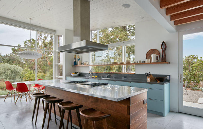 In this contemporary kitchen there's a large island with a walnut base and a stainless steel countertop that surrounds the cooktop. Against the wall, light blue cabinets add a pop of color and an open shelf lets the home owners display their favorite kitchen items.