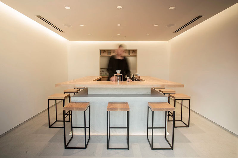 The interior of Tokyo Saryo features a simple bar that sits centered on a concrete floor and is surrounded by stools made from black steel frames and wooden seats. The walls have been left bare and a drop ceiling holds both hidden lighting and six small pot lights to create a soft glow throughout the space.
